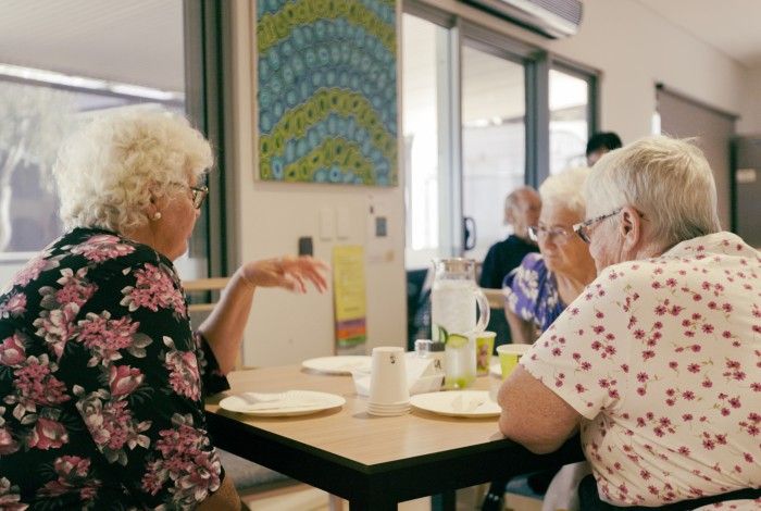 Three elderly women sit at a table in a communal dining area, engaged in conversation with plates, cups, and a water jug in front of them.