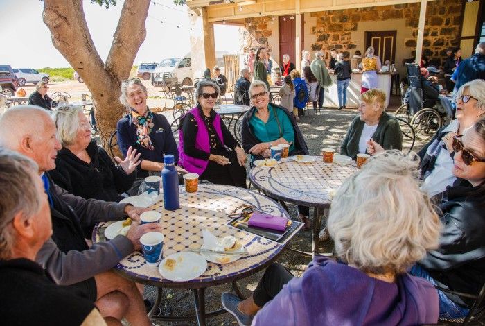 A group of older adults sit together at outdoor café tables beneath a large tree, enjoying refreshments and conversation at a social gathering. Additional attendees are seated and mingling around a historic stone building in the background on a sunny day.