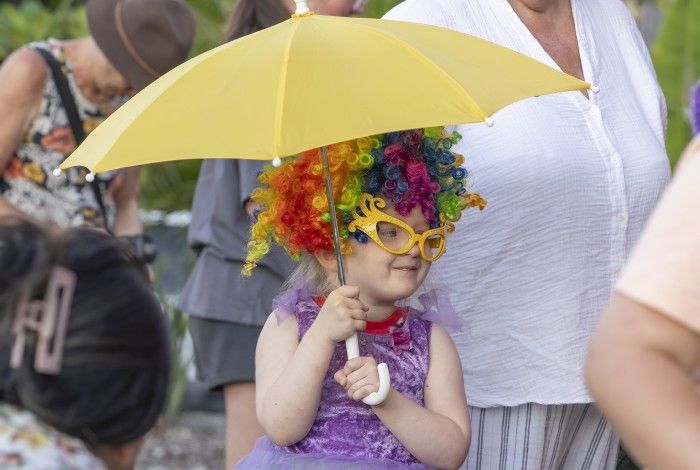 A young child wearing a rainbow curly wig, oversized yellow novelty glasses and a purple dress smiles while holding a bright yellow umbrella. The child is surrounded by adults at an outdoor community event, creating a colourful and playful scene.