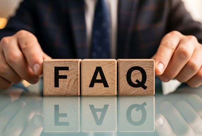 Close-up of three wooden blocks on a reflective desk spelling “FAQ” in bold black letters, positioned between a person's hands. The person is wearing a dark business jacket, and the softly blurred background creates a professional office setting. The blocks and their reflection are in sharp focus.
