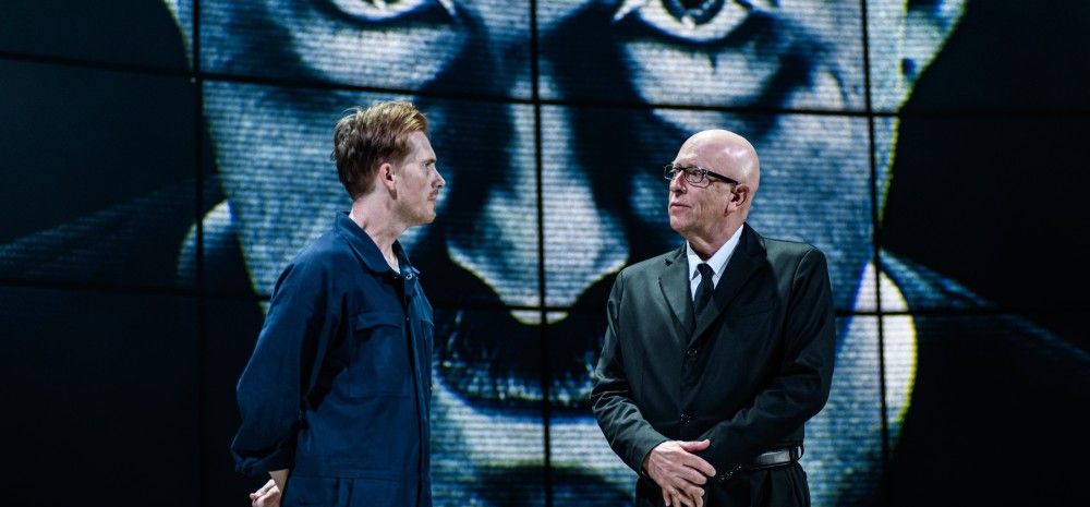 Two men stand facing each other, one in a jumpsuit and one in a suit. Behind them, a massive projected image of a face with penetrating eyes looms, giving a sense of authority and control.